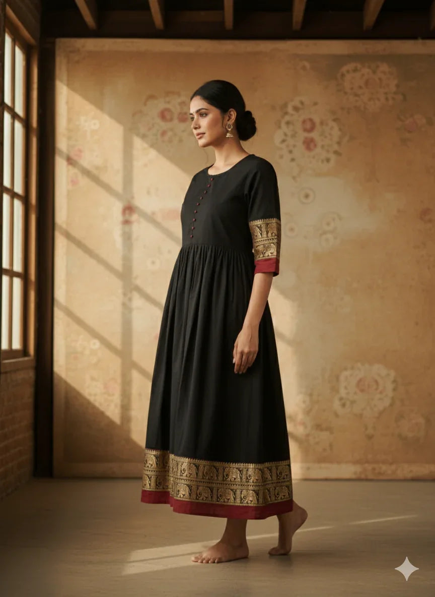 Woman in a black dress with gold and red details standing in a room with floral wallpaper.